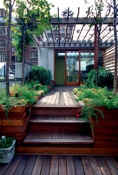 Modern rooftop deck with wooden stairs, planters, and glass structure overlooking brick buildings.