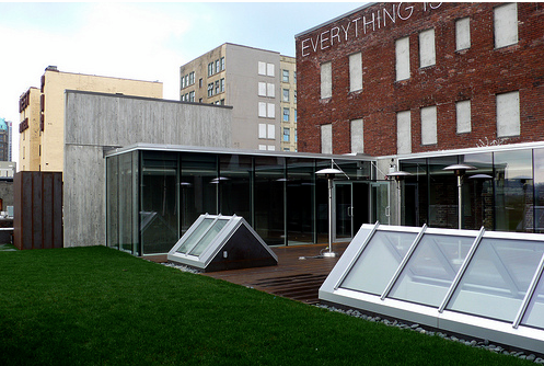 Modern glass and concrete pavilion on green roof deck with skylight windows and urban brick buildings backdrop