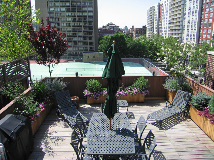 Urban rooftop garden with dining furniture, flowering plants, and green umbrella overlooking city skyline.