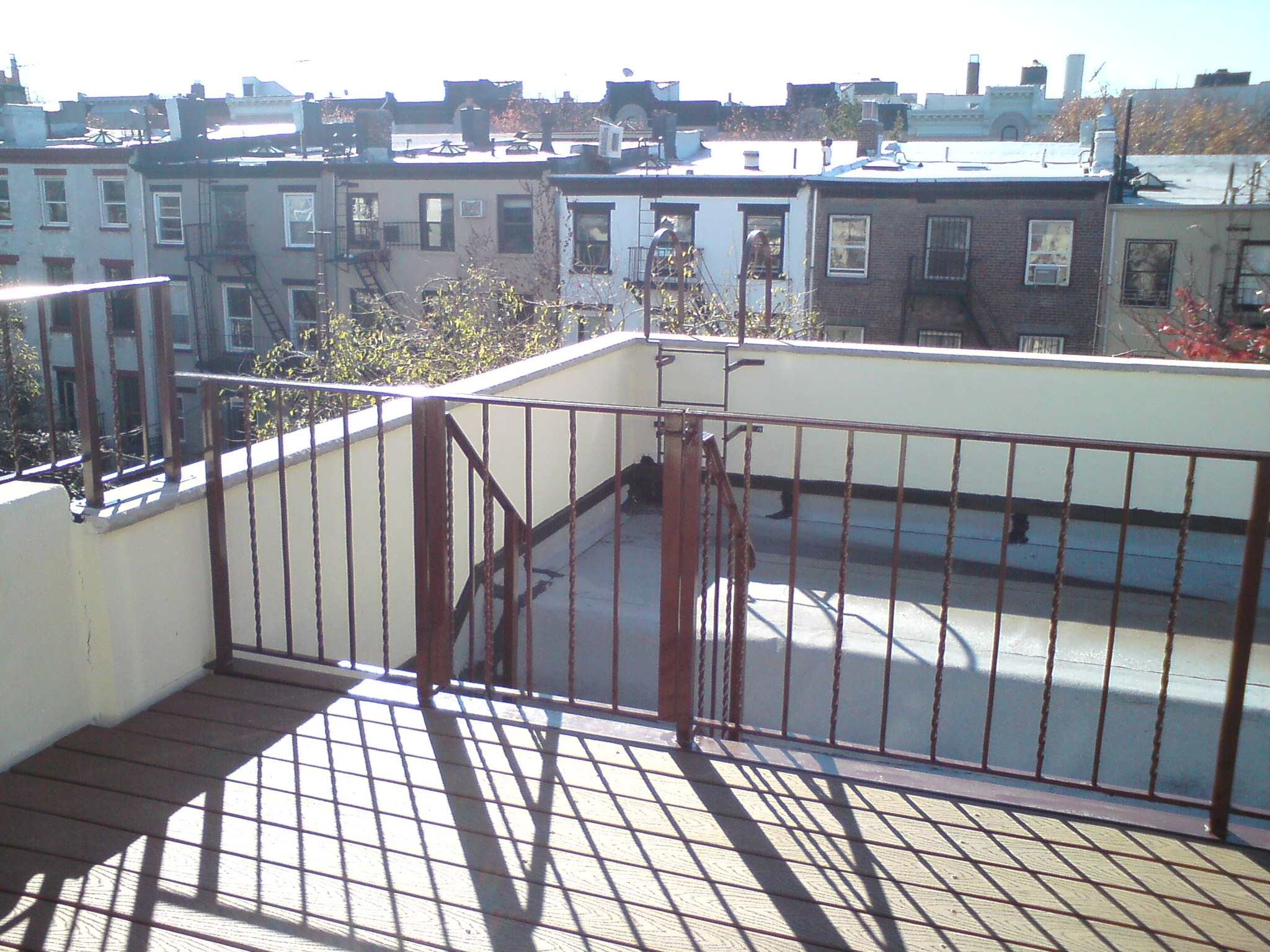 Sunny rooftop deck with metal railing overlooking neighboring brick buildings and water.