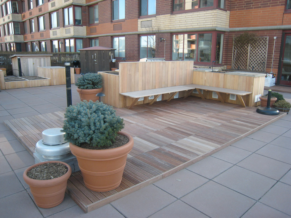 Modern rooftop deck with wooden L-shaped bench seating and potted plants overlooking brick buildings.