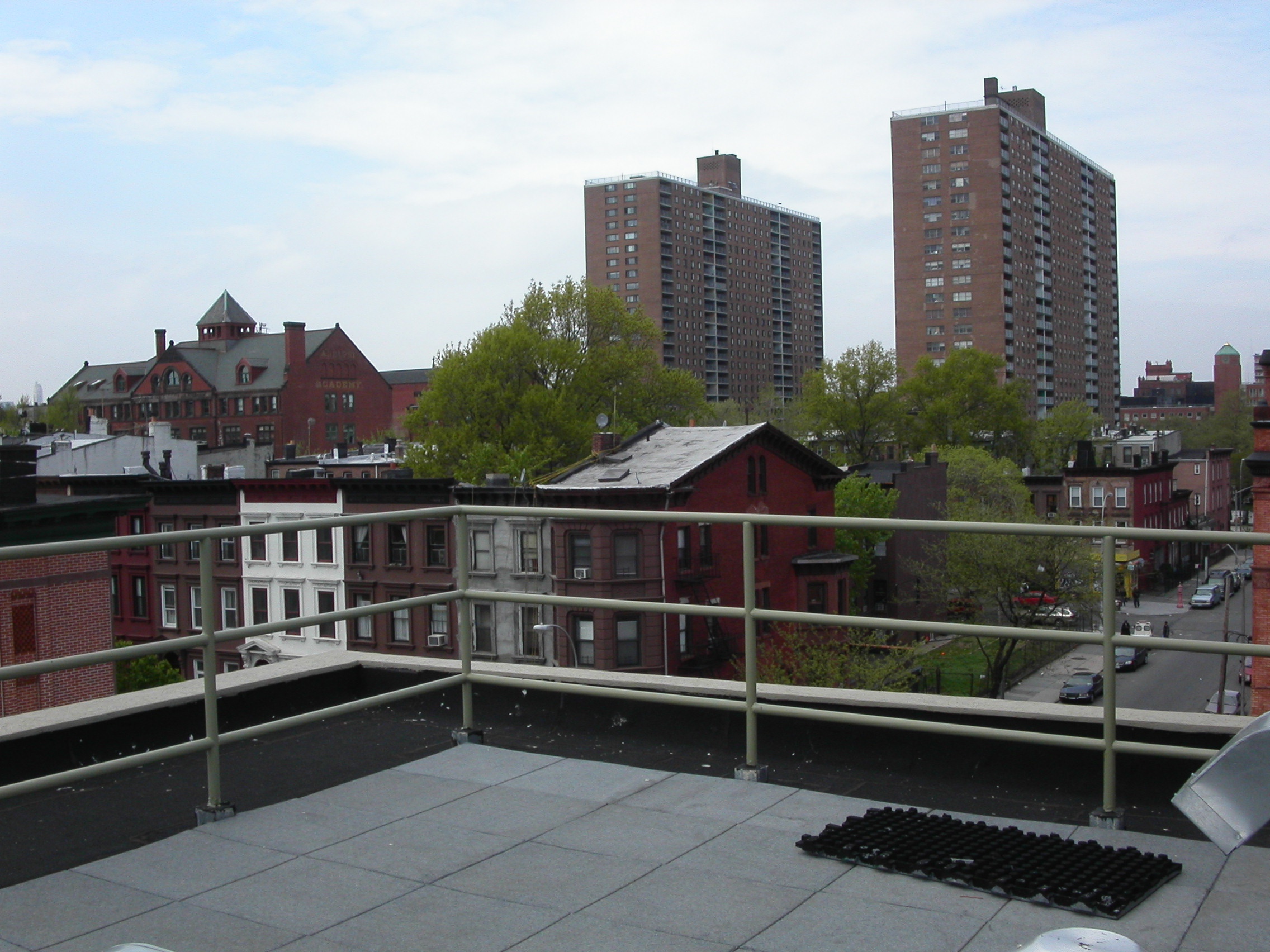 Rooftop deck with metal railing overlooking brick buildings and high-rise apartments in Brooklyn