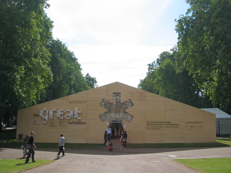 Tan pavilion building with 'the great exhibition' text and symmetrical architectural motif, tree-lined approach.