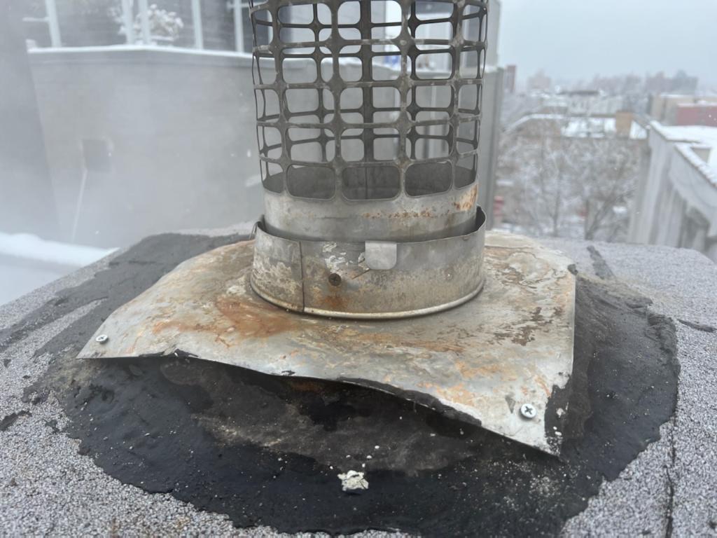 Corroded metal boiler vent with grid cap mounted on flat roof, showing rust and wear.