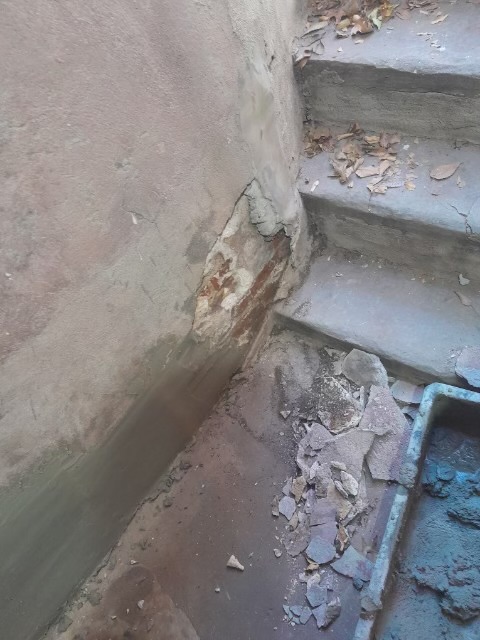 Deteriorated concrete wall corner with peeling plaster, exposed aggregate, and basement stairs visible