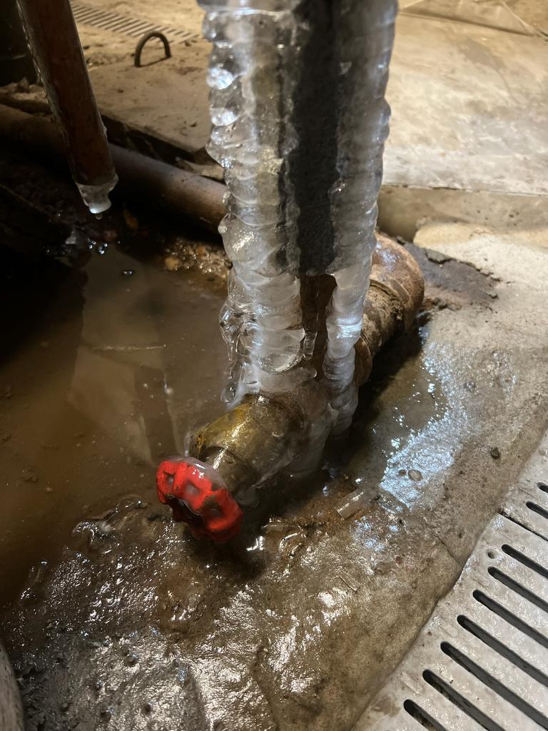 Frozen water pipe with ice buildup and red valve, surrounded by water pooling on concrete floor.